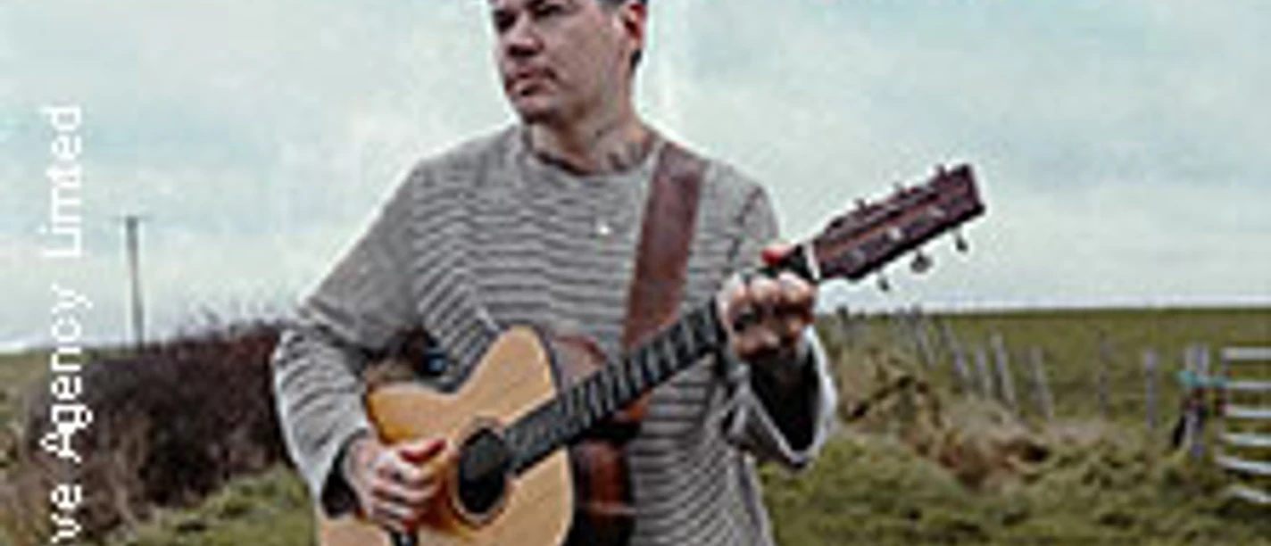 Person mit Gitarre auf einer Landstraße unter bewölktem Himmel.