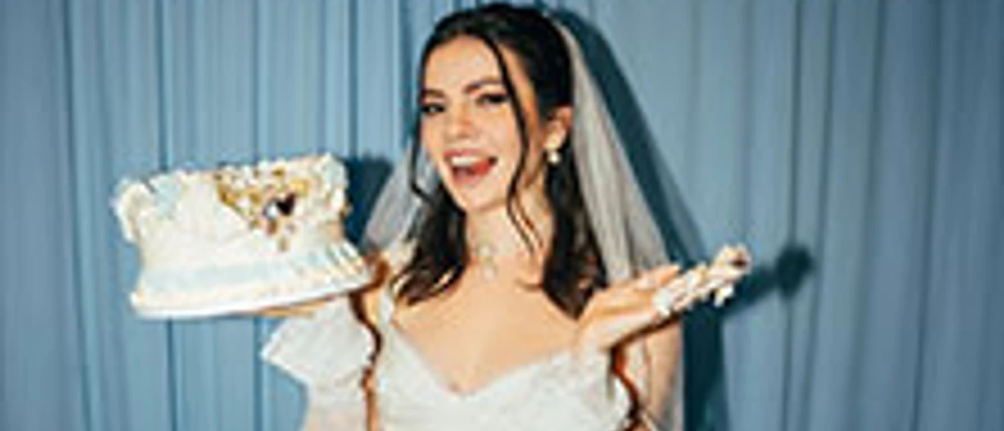 Frau im Brautkleid lächelnd vor blauem Vorhang, hält angeschnittene Hochzeitstorte