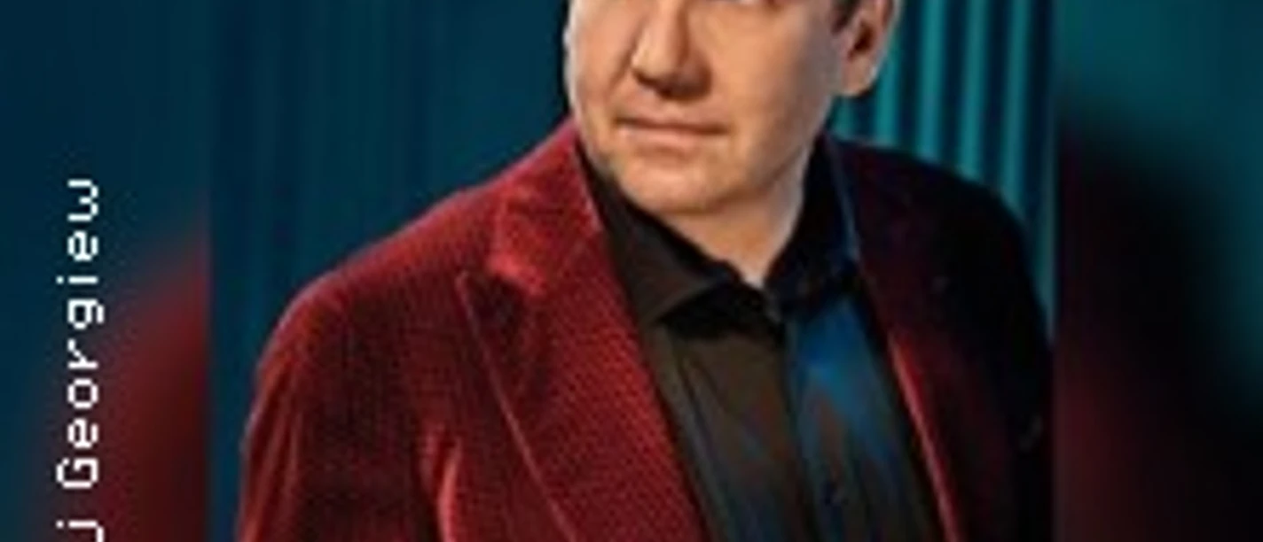 Mann im roten Sakko vor blauem Vorhang, blickt seitlich mit ruhiger Haltung.