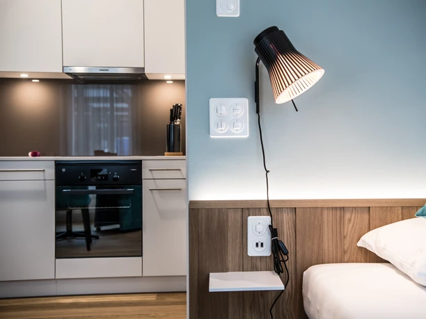 Aparthotel Luzern West Alberswil Detailaufnahme im Aparthotel Luzern West mit Schlafzimmerbereich, Wandlampe und Blick zur modernen Küche