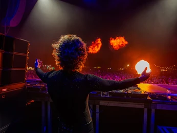 Rainbow Festival DJ-Person auf Bühne mit Feuer-Effekten über einer großen, farbig beleuchteten Festivalmenge.DJ person on stage with fire effects above a large, colorfully illuminated festival crowd.