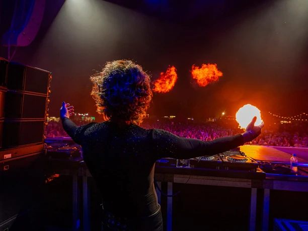 Rainbow Festival DJ person on stage with fire effects above a large, colorfully illuminated festival crowd.