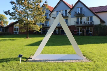 Skulpturenpark -Weiß Gefaltet Weiße dreieckige Skulptur auf grüner Wiese vor modernen Gebäuden im sonnigen Skulpturenpark Otterndorf.