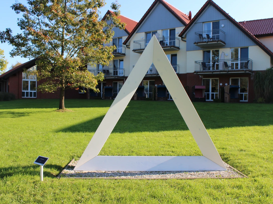 Skulpturenpark -Weiß Gefaltet Weiße dreieckige Skulptur auf grüner Wiese vor modernen Gebäuden im sonnigen Skulpturenpark Otterndorf.