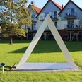 Skulpturenpark -Weiß Gefaltet Weiße dreieckige Skulptur auf grüner Wiese vor modernen Gebäuden im sonnigen Skulpturenpark Otterndorf.