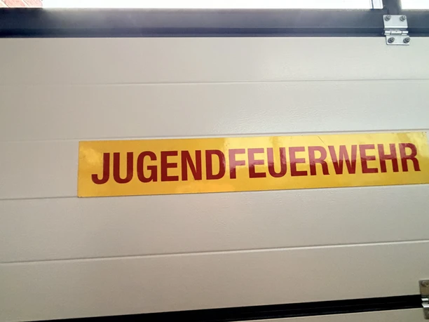 IMG_0805.jpg Gelbes Schild mit roter Aufschrift Jugendfeuerwehr an einer hellen Garagentür der Feuerwehr.