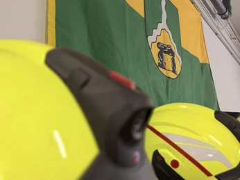 IMG_0812.jpg Reihe leuchtend gelber Feuerwehrhelme vor einer grünen Flagge mit gelbem Wappen an der Wand.