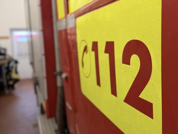 IMG_0814.jpg Rotes Feuerwehrfahrzeug mit gelbem Notrufzeichen 112, im Hintergrund Feuerwehrkleidung im Raum.