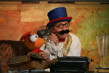 Dolittle.jpg Person mit blauem Zylinderhut und roter Fliege sitzt vor einer alten Landkarte neben einer Vogelpuppe und einer Schreibmaschine