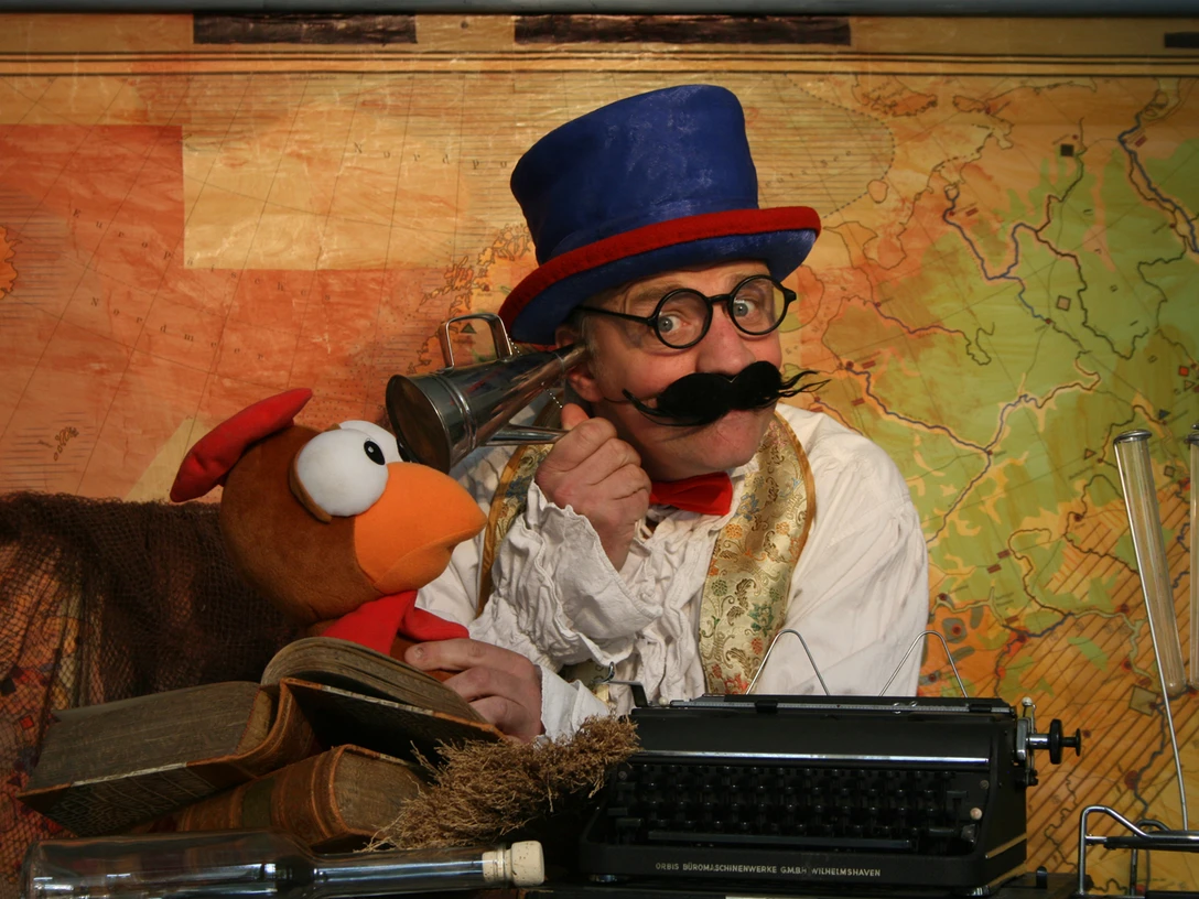 Dolittle.jpg Person mit blauem Zylinderhut und roter Fliege sitzt vor einer alten Landkarte neben einer Vogelpuppe und einer Schreibmaschine