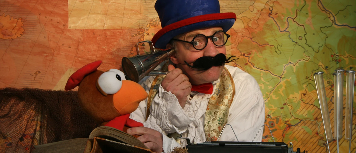 Dolittle.jpg Person mit blauem Zylinderhut und roter Fliege sitzt vor einer alten Landkarte neben einer Vogelpuppe und einer Schreibmaschine