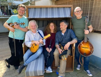 Ceann Mara.jpg Fünf Personen mit Musikinstrumenten sitzen und stehen um eine Strandkorbbank auf gepflastertem Boden.