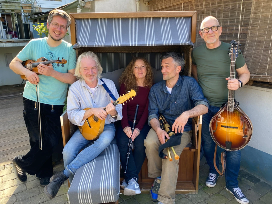 Ceann Mara.jpg Fünf Personen mit Musikinstrumenten sitzen und stehen um eine Strandkorbbank auf gepflastertem Boden.