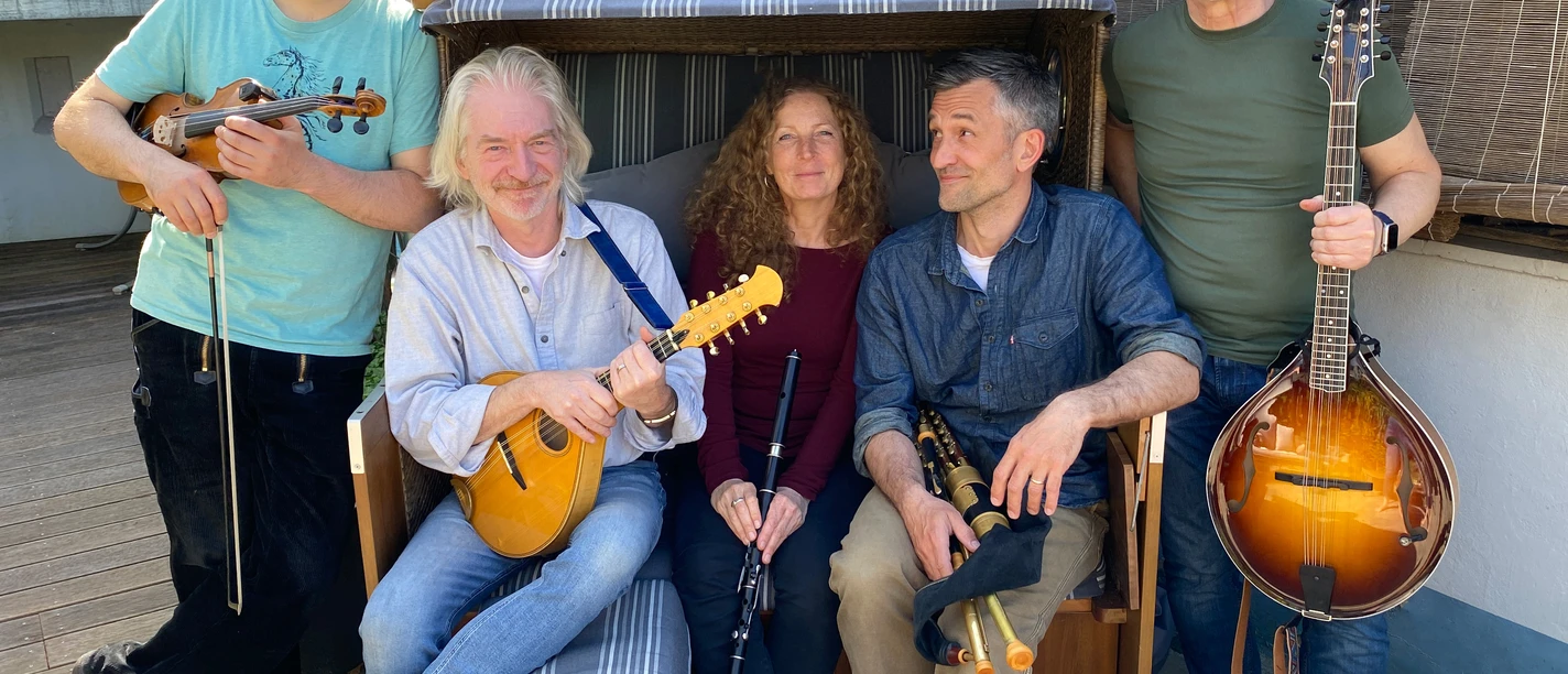 Ceann Mara.jpg Fünf Personen mit Musikinstrumenten sitzen und stehen um eine Strandkorbbank auf gepflastertem Boden.