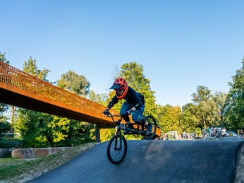 Kind mit Helm fährt BMX über Wellen des Pumptracks, im Hintergrund Fußgängerbrücke und Zuschauer.