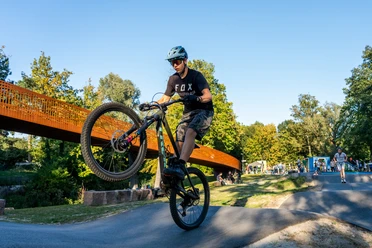 Mountainbiker hebt Vorderrad auf Pumptrack, weitere Fahrer und Fußgänger im Hintergrund