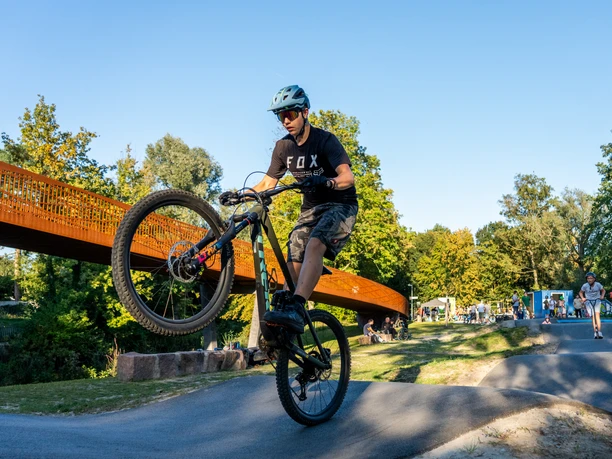 Area61 Mountainbiker hebt Vorderrad auf Pumptrack, weitere Fahrer und Fußgänger im Hintergrund