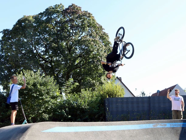 BMX-Fahrer dreht einen Rückwärtssalto über dem Pumptrack, zwei Zuschauer filmen am Rand.