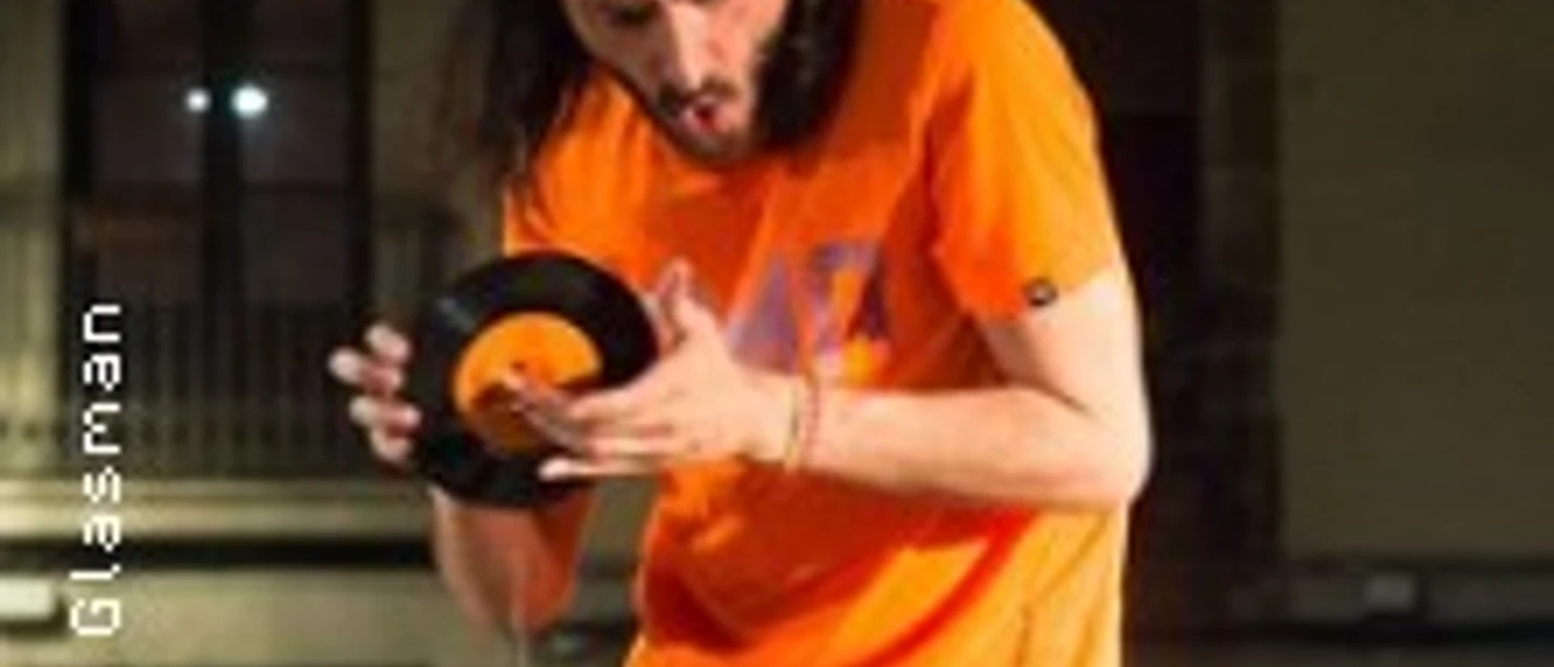 Ein DJ mit Kopfhörern hält eine kleine Schallplatte über einem beleuchteten Mischpult.