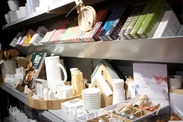 2026-01-Dodt-Schreibwaren-geschenke.jpg Regal mit farbigen Notizbüchern, Geschenkartikeln, Keramikgeschirr und Wohnaccessoires in hellem Licht.
