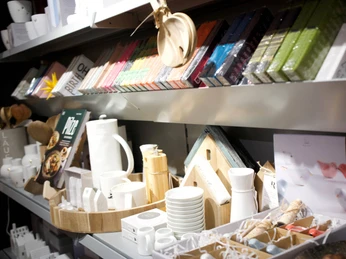 2026-01-Dodt-Schreibwaren-geschenke.jpg Regal mit farbigen Notizbüchern, Geschenkartikeln, Keramikgeschirr und Wohnaccessoires in hellem Licht.