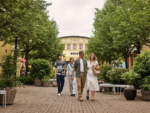 Wertheim Village -Eine Gruppe von Personen im Wertheim Village“ Eine Gruppe von Personen spaziert entspannt durch die begrünte Einkaufspassage von Wertheim Village.