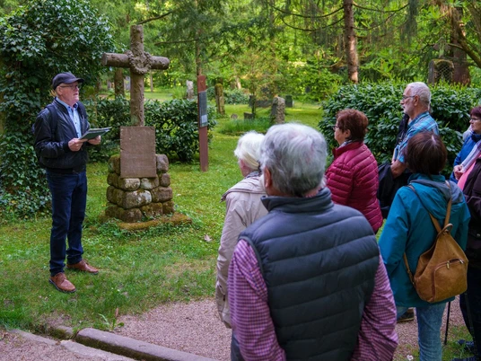 Gruppe hört dem Gästeführer zu, der vor einem Steinkreuz seinen Vortrag hält