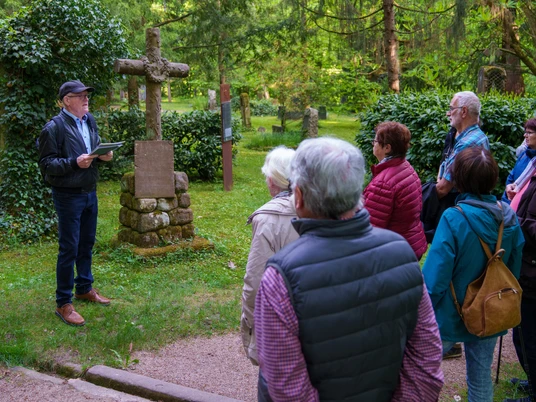 Gruppe hört dem Gästeführer zu, der vor einem Steinkreuz seinen Vortrag hält
