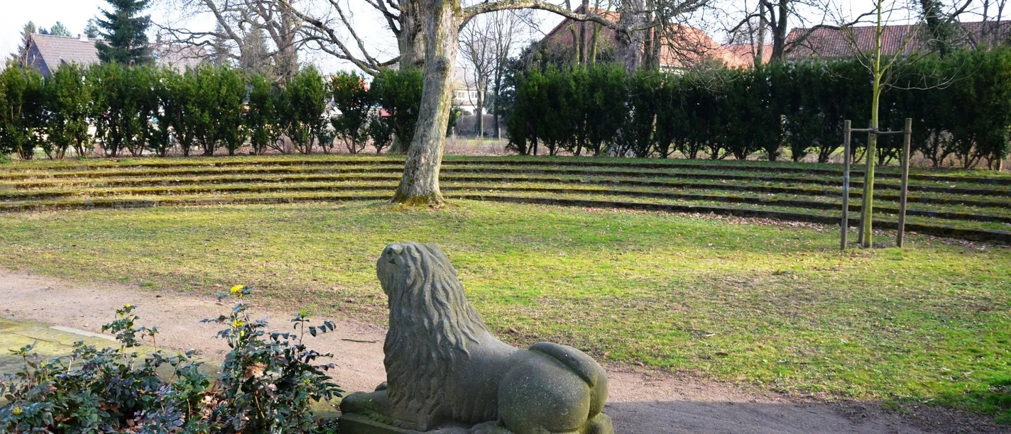 Salzgitter-Flachstoeckheim_Freilichtbuehne_HD_16zu9.JPG Steinerne Löwenfigur vor halbrunder Freilichtbühne mit Rasenstufen und alten Bäumen im Gutspark.