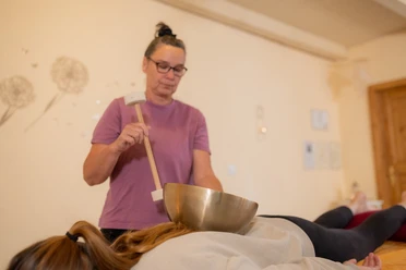 Klangmassagen Spiegelhaus Woltzeten Frau führt Klangmassage mit Klangschale auf dem Rücken einer liegenden Person in ruhigem Raum durch.