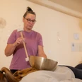 Klangmassagen Spiegelhaus Woltzeten Frau führt Klangmassage mit Klangschale auf dem Rücken einer liegenden Person in ruhigem Raum durch.