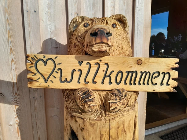 Herzlich Willkommen in der Atzmannhütte Geschnitzter Holzbär mit Schild "Willkommen" in den Pfoten