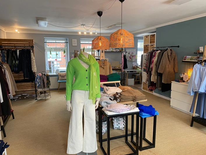 Cindys-frauenzimmer-bispingen-innenraum.jpg Innenraum eines Modegeschäfts mit hellen Wänden, Kleiderständern, Tisch mit Kleidung und Schaufensterpuppe.