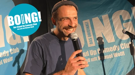 BOING! Comedy Club Köln Lachender Comedian mit Mikrofon auf Bühne vor blauem BOING!-Hintergrund.