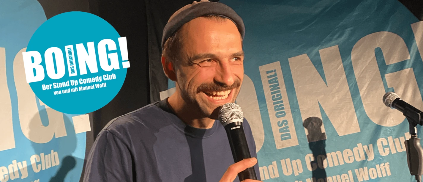BOING! Comedy Club Köln Lachender Comedian mit Mikrofon auf Bühne vor blauem BOING!-Hintergrund.
