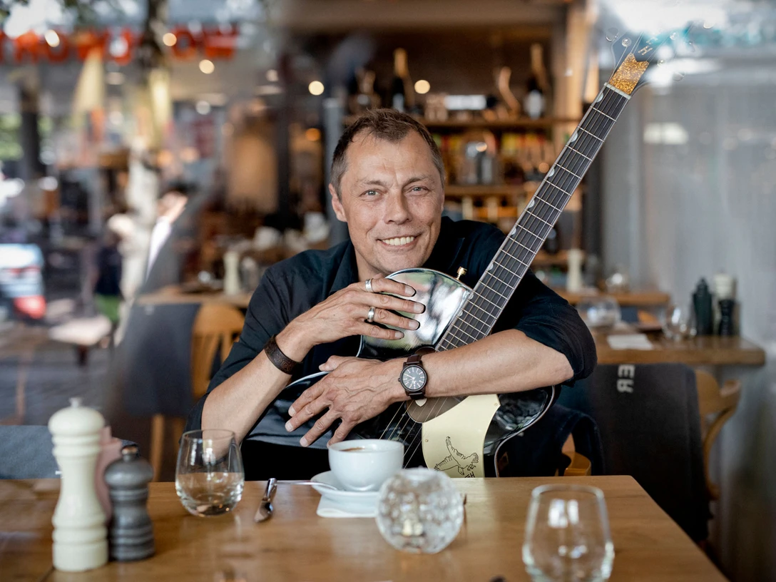 buesum-Georgie Carbutler-2560x1707.jpeg Ein Mann sitzt im Restaurant an einem Tisch mit einem Kaffee vor sich auf dem Tisch. Er hält eine Gitarre im Arm.