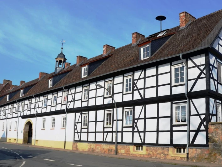 Gutshof_Flachstöckheim (2)_HD_16zu9.JPG Langgestrecktes Fachwerkgebäude mit Torbogen, dunklem Gebälk und roten Ziegeldächern unter blauem Himmel.