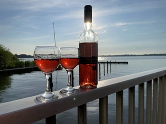 Roséwein und zwei Gläser bei tiefstehender Sonne am Meer Zwei Gläser Roséwein und eine Flasche auf einem Geländer mit Blick auf den See im Abendlicht