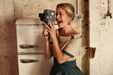 Sabrina - Schlagershow-2560x1707 .jpg Eine Frau mit Flechtfrisur und Kleid vor einem alten Kühlschrank. Sie hält eine alte Filmkamera in der Hand, durch die sie durchschaut.