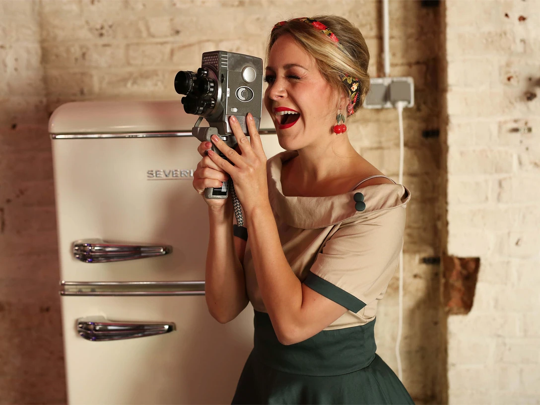 Sabrina - Schlagershow-2560x1707 .jpg Eine Frau mit Flechtfrisur und Kleid vor einem alten Kühlschrank. Sie hält eine alte Filmkamera in der Hand, durch die sie durchschaut.