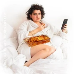 Person im Bademantel sitzt im Bett, hält ein Smartphone und hat eine Auflaufform mit Lasagne auf dem Schoß.