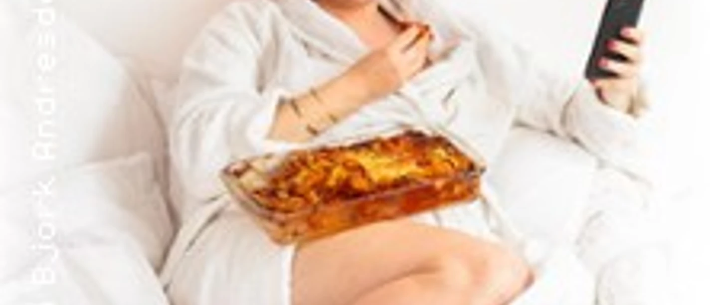 Person im Bademantel sitzt im Bett, hält ein Smartphone und hat eine Auflaufform mit Lasagne auf dem Schoß.