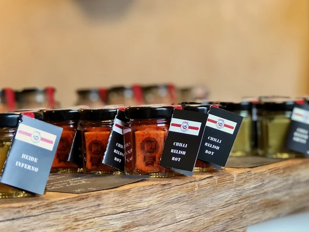 Laden-Chilli-Manufaktur-Susanne-Menke-Luenburger-Heide-1.jpeg Reihe bunter Chutney- und Relishgläser mit schwarzen Etiketten auf einer Holztheke im Hofladen.