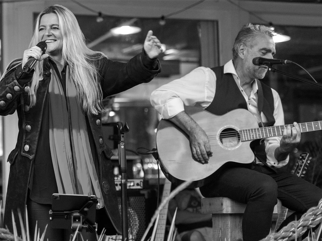 Pressebild Tinas Livingroom 2560x1707.jpg Eine Sängerin steht auf der Bühne. Daneben sitzt auf einem Hocker ein Mann, der Gitarre spielt.