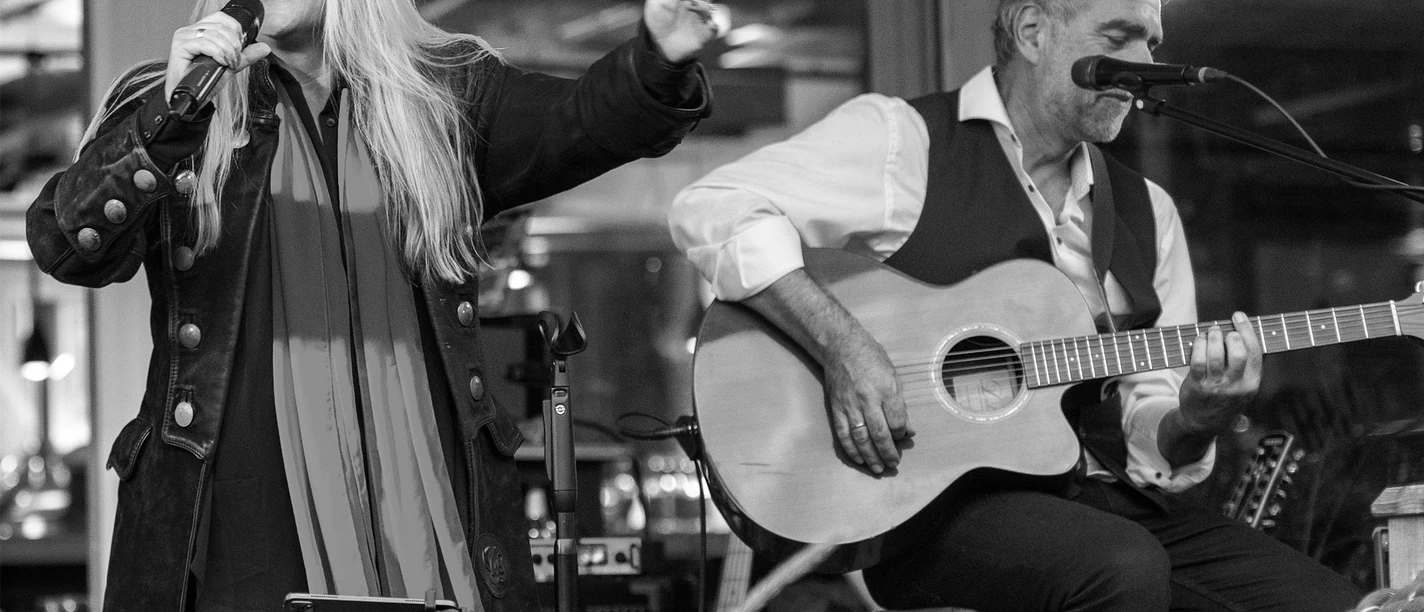 Pressebild Tinas Livingroom 2560x1707.jpg Eine Sängerin steht auf der Bühne. Daneben sitzt auf einem Hocker ein Mann, der Gitarre spielt.