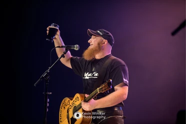 Mac Piet in Büsum Ein Mann mit einer Cap und einem schwarzen T-Shirt hält eine Gitarre in der Hand und hebt mit einer Hand ein Bierglas in die Luft. Vor ihm steht ein Mikrofonständer mit Mikrofon.