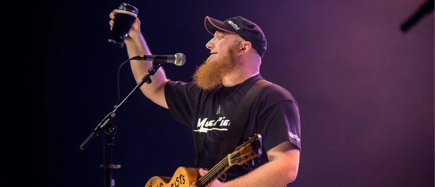 Mac Piet in Büsum Ein Mann mit einer Cap und einem schwarzen T-Shirt hält eine Gitarre in der Hand und hebt mit einer Hand ein Bierglas in die Luft. Vor ihm steht ein Mikrofonständer mit Mikrofon.
