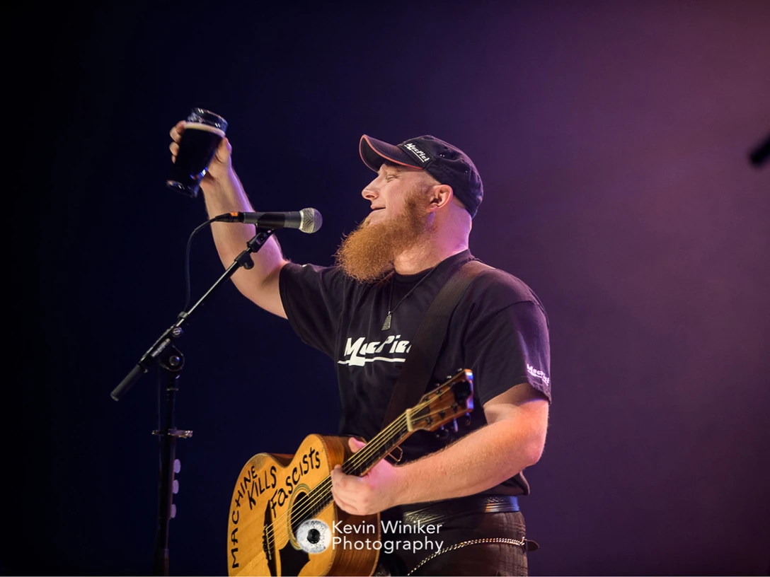 Pressefoto_Mac Piet_Fotocredit_Kevin Winiker-2560x1707.jpeg Ein Mann mit einer Cap und einem schwarzen T-Shirt hält eine Gitarre in der Hand und hebt mit einer Hand ein Bierglas in die Luft. Vor ihm steht ein Mikrofonständer mit Mikrofon.