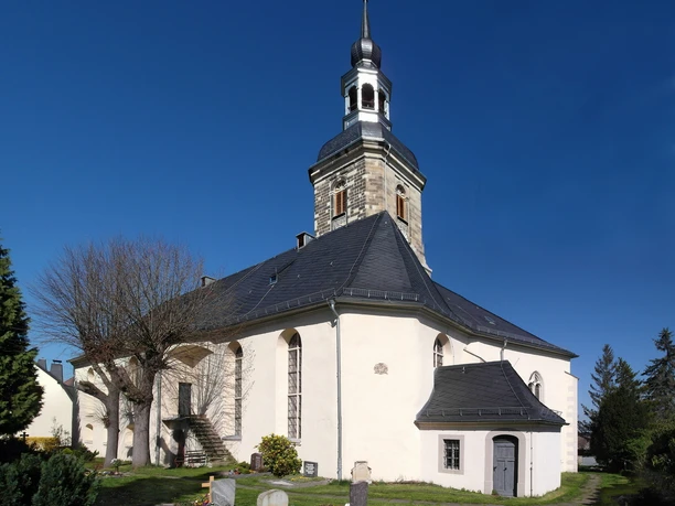 Weißes Kirchengebäude mit schwarzem Dach und spitzem Turm, umgeben von Gräbern und Bäumen.