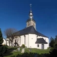 Kirche in Burkhardswalde Weißes Kirchengebäude mit schwarzem Dach und spitzem Turm, umgeben von Gräbern und Bäumen.
